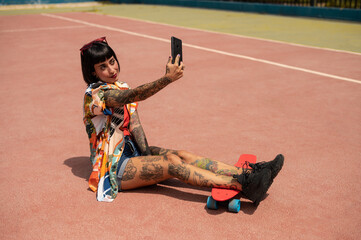Atractiva mujer con tatuajes en el cuerpo haciendose unas fotos con la camara del telefono en una pista deportiva con un skate