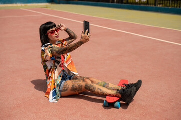 Caucasian woman with tattoos and short hair on a sports court with a skateboard using her phone's camera