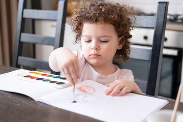 curly child girl painting with watercolor paints at home at kitchen table