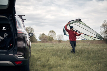 Windsurfer unpacking equipment from a car in nature near the lake shore.