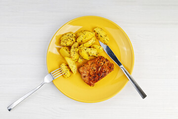 Grilled turkey steak with a garnish of baked potatoes in a yellow plate on white wooden table. Top view.