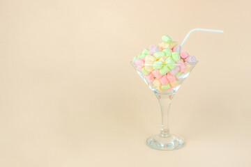 Brignt marshmallows for sweet break in wineglass with straw on pastel beige background.
