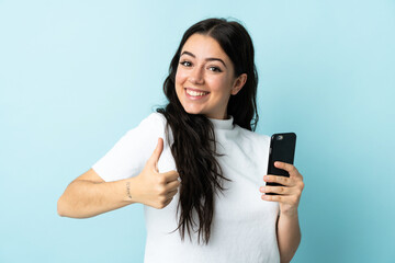 Young woman using mobile phone isolated on blue background with thumbs up because something good has happened
