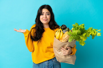 Young woman holding a grocery shopping bag isolated on blue background having doubts while raising...