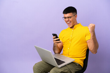 Young man sitting on a chair with laptop with phone in victory position