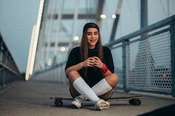Young woman sitting on her longboard and using a smartphone while on the bridge