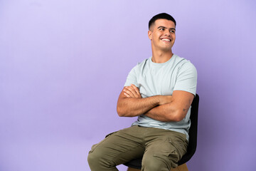 Young man sitting on a chair over isolated background with arms crossed and happy