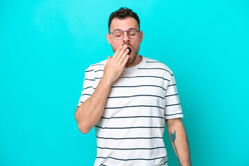 Young Brazilian man isolated on blue background yawning and covering wide open mouth with hand