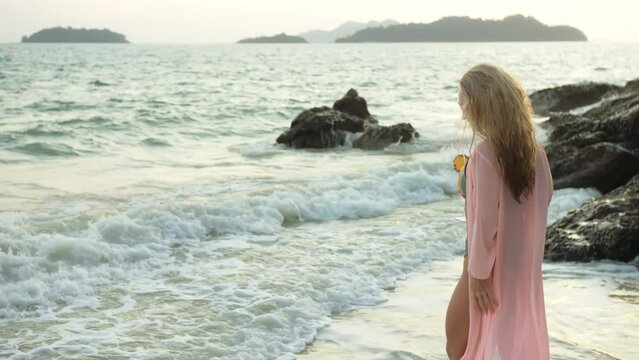 Attractive woman stand on a reef rock stone in sea on golden sunset. Girl on tropical beach in green swimsuit and flutter in wind pink tunic silk shirt cape, drinks her orange cocktail Pina Colada