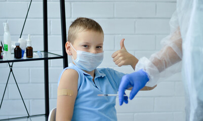 Close-up of a boy in a blue T-shirt and a medical mask, who raised his finger up after vaccination. A doctor in gloves with a syringe.