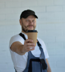 Portrait of young handsome caucasian man in a white T-shirt and overalls, in a black cap holding cup of coffee to go.
