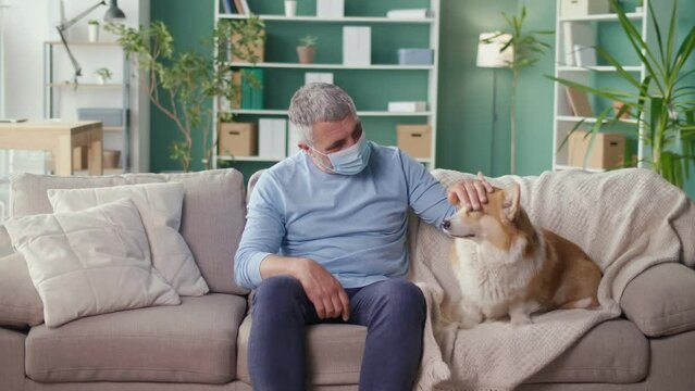 A Sad Lonely Elderly Man in a Medical Mask Stroking His Dog While Sitting on the Couch at Home. Canistherapy, Best Friend, Help, Pet, Mental Health. Pandemic, Social Distancing and Self-isolation.
