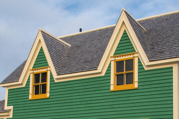 Two small single hung windows under gable or triangle shaped dormers on a vintage green exterior building wall with wood casement yellow trim. There's a blue sky with white clouds in the background