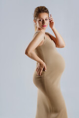 Fototapeta premium Studio portrait of an elegant young adult pregnant woman in a beige dress on a gray background, the concept of a happy pregnancy.