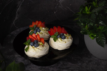 Set of Pavlova desserts on a dark surface, topped with a mix of berries and set beside a green plant.