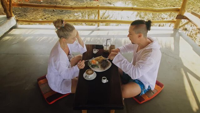 Attractive young man and woman eat and drink tasty beverages spending time in traditional local floating cafe on water. Loving happy couple having breakfast outdoors, feed each other. Tropical view