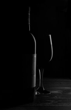 Black and white dark photography, a bottle of wine and a glass cup on a rustic table, lit by a rim light, dark or black ambient, merlot cabernet sauvignon fine art abstract