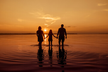 Full body silhouette of an unrecognizable family holding hands and walking with their backs to the camera on the water of a river, during sunset, copy space, toned image