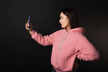 Cute young brunette in a pink hoodie uses a smartphone to take a selfie, portrait on a black background