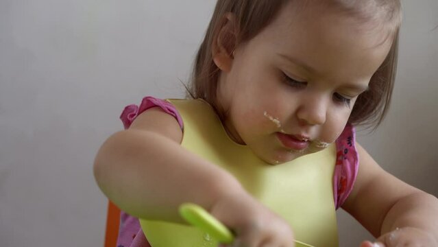 Close up of lovely baby girl eating have breakfast eggs vegetables sausage by herself at home. Adorable possitive happy toddler child eats with dirty face at table indoor. Food, nutrition healthy life