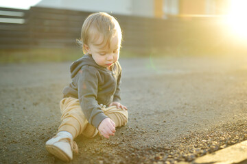 Funny toddler boy sitting on the ground outdoors on sunny summer day. Child exploring nature.