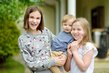 Two big sisters and their toddler brother having fun outdoors. Two young girls holding baby boy on summer day. Children with large age gap. Big age difference between siblings.