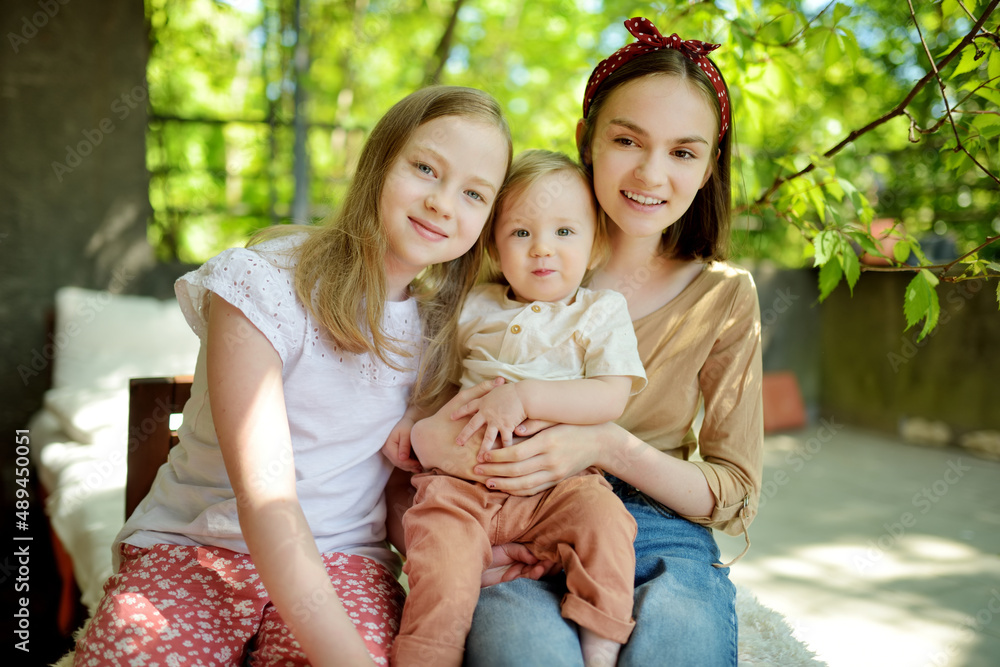 Wall mural Two big sisters and their toddler brother having fun outdoors. Two young girls holding baby boy on summer day. Children with large age gap. Big age difference between siblings.