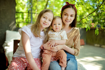 Two big sisters and their toddler brother having fun outdoors. Two young girls holding baby boy on summer day. Children with large age gap. Big age difference between siblings.