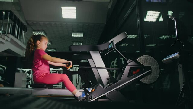 Fit Athletic Baby Girl Work out on Rowing Machine, Doing Her Fitness Exercise. Children Activity Training in Modern Gym. Sports Child Workout in Exercising on Row Machine Fitness Center.