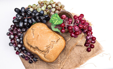 A loaf of coarse rye homemade bread with a cracked toasted crust and bunches of red, black and green grapes lying on a burlap. Flesh and Blood of Jesus Christ