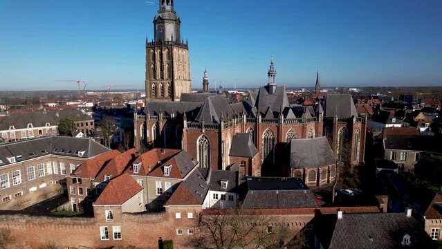 Sideways aerial movement following the medieval city wall of tower town Zutphen with authentic historic rooftops and buildings with Walburgiskerk church towering over against a clear blue sky.