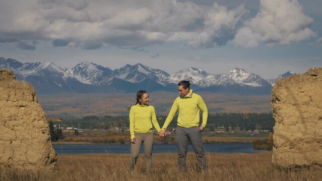 Man and woman in yellow green sportswear. Lovely couple of travelers hug and kiss near old stone enjoying highland landscape. Two travelers are walking against the backdrop of snow-capped mountains.