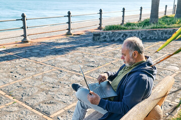 Handsome bearded mature man with laptop computer working outdoors while sitting on bench at the seaside. Modern lifestyle, connection, business, freelance work concept.