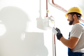 Low angle view of technician, heating fitter in hard hat and protective gloves making notes while checking gas consumption at gas pipe and meter counter - obrazy, fototapety, plakaty