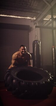 A Vertical Video Of A Hispanic Bodybuilder Tyre Flipping In An MMA Gym. Shot In A Crossfit Boxing Gym With Low Key Lighting And A Scattering Of Haze. Captured On Red Digital Cinema Camera 