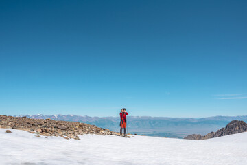 Fototapeta na wymiar Tourist in red walks on snow mountain near abyss edge on high altitude under blue sky in sunny day. Man with camera on snowy mountain near precipice edge with view to large mountain range in away.