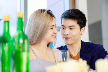 Obraz na płótnie Canvas Millennial young lover couple Asian husband in formal suit and beautiful wife smiling holding tall glasses wine toasting cheers together in happy new year party at decorated beverage and food table