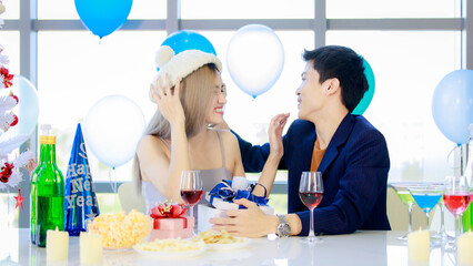 Millennial young lover couple Asian husband in formal suit and beautiful wife smiling holding tall glasses wine toasting cheers together in happy new year party at decorated beverage and food table