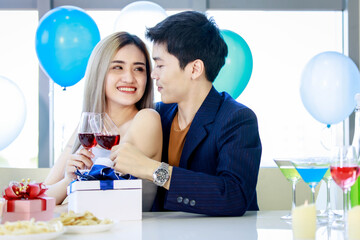 Millennial young lover couple Asian husband in formal suit and beautiful wife smiling holding tall glasses wine toasting cheers together in happy new year party at decorated beverage and food table