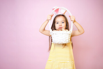 Happy Easter. A cute funny girl with bunny ears holds a basket of chocolate eggs in her hands. Pink background.