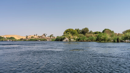 Fototapeta na wymiar Lush green vegetation, picturesque boulders, sand dunes are visible on the banks of the Nile. Ripples on the blue water. Clear azure sky. A sunny day. Egypt.