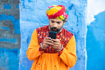 Surprised traditional Indian man wearing colorful outfits and turban using smart phone,Amazed rural...