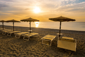  empty beach during beautiful sunrise or sunset with chaise loungues and nice umbrellas with blue sea, sun glow and amazing cloudy sky on thr background