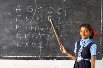 girl kid from blackboard at public school teaching alphabets to classmates by looking at camera -...