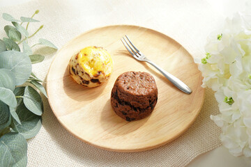 Two Sconces, chocolate and raisins flavor, serving on wooden plate and forks placed on placemat and decorate with white flower and green leaves in morning light. coffee break or tea Time. Flat lay.