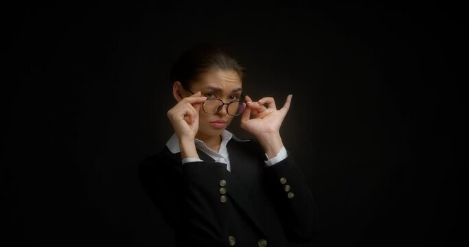 Business woman in perplexity lowers her glasses and looks at the camera with a dissatisfied expression on her face. Serious Asian woman is not satisfied with the answer. Isolated on a black background