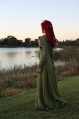 Full length portrait of red haired woman wearing a  beautiful  green medieval fantasy gown. Posing with gestural hands on a enchanted forest background.