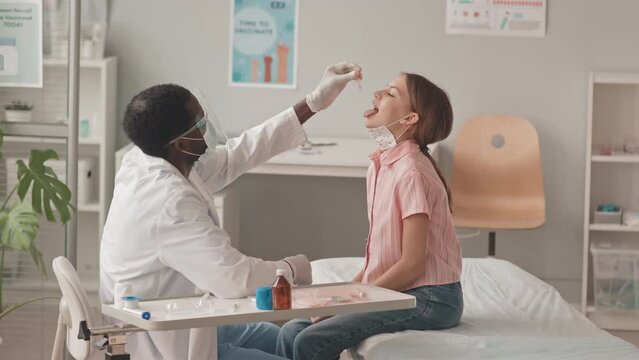 Medium slowmo shot of 13-year-old Caucasian girl getting oral vaccination from young African-American doctor in face shield