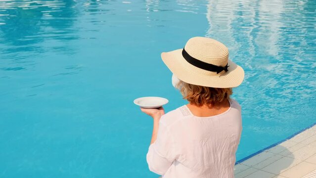 female in a straw hat wearing a white dress relaxing by the pool with a cup of coffee, vacation concept, good morning. woman sitting by the pool