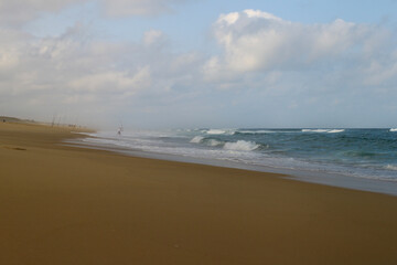 The fisherman in action at the beach. A fisherman throws his feet into the waves. The sandy beach continues very far. The sky is overcast, the waves of the ocean are breaking towards the beach.
